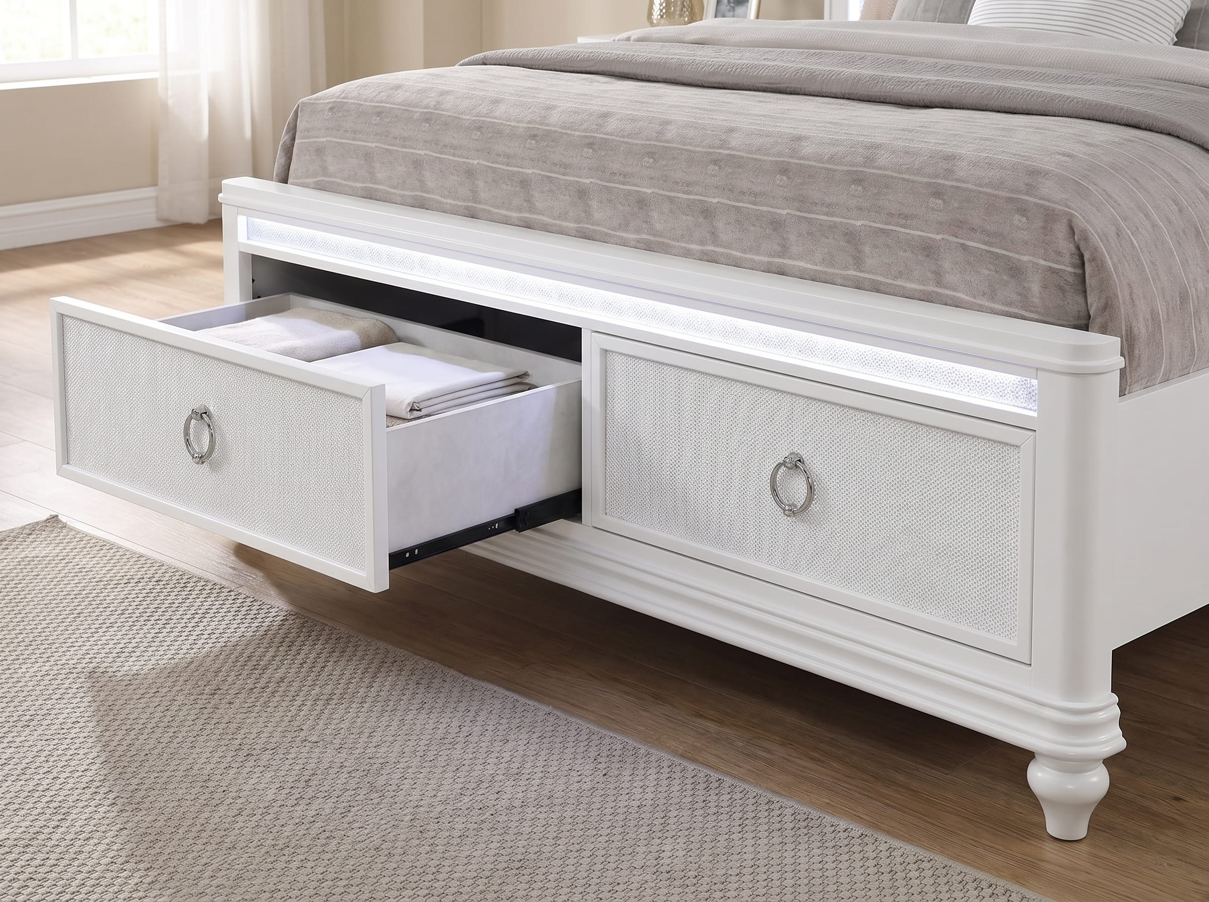 Abigale white storage bed footboard with one drawer slid open on its runners, showing velvet-lined interior with folded linens, the second drawer closed, and the integrated LED light channel lit across the footboard rail
